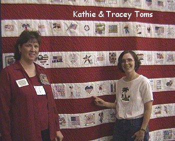 The project organizer and Tracey Toms in front of a quilt, one pointing to a square